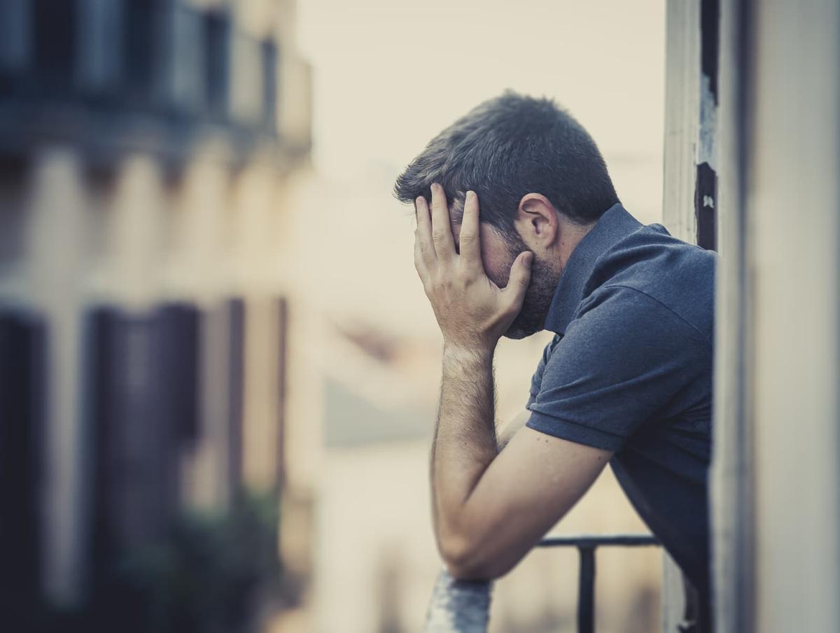 Man standing on a balcony, hunched over, holding his head in his hands.