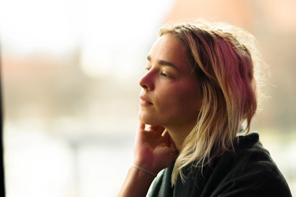 Woman with blonde hair looking out a window with a thoughtful expression, gently touching her neck