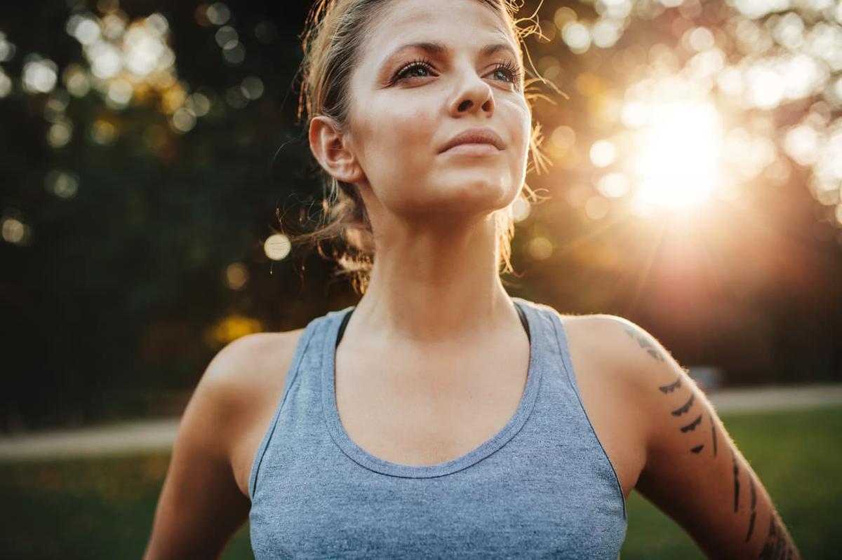 A woman in a grey tank top stands outdoors during golden hour, looking thoughtfully into the distance with a sun flare in the background