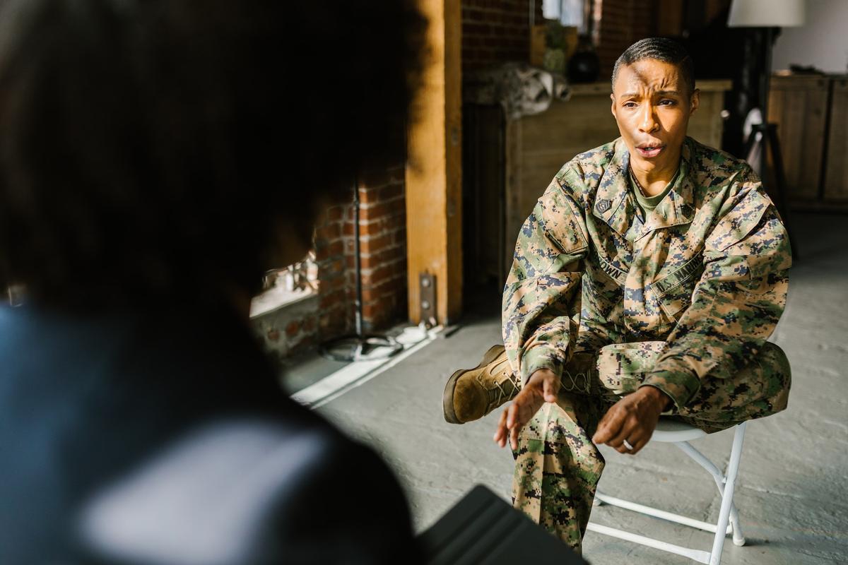A service member in camouflage uniform sits indoors and speaks earnestly to another person, suggesting a serious conversation about mental health, substance use, or recovery support.