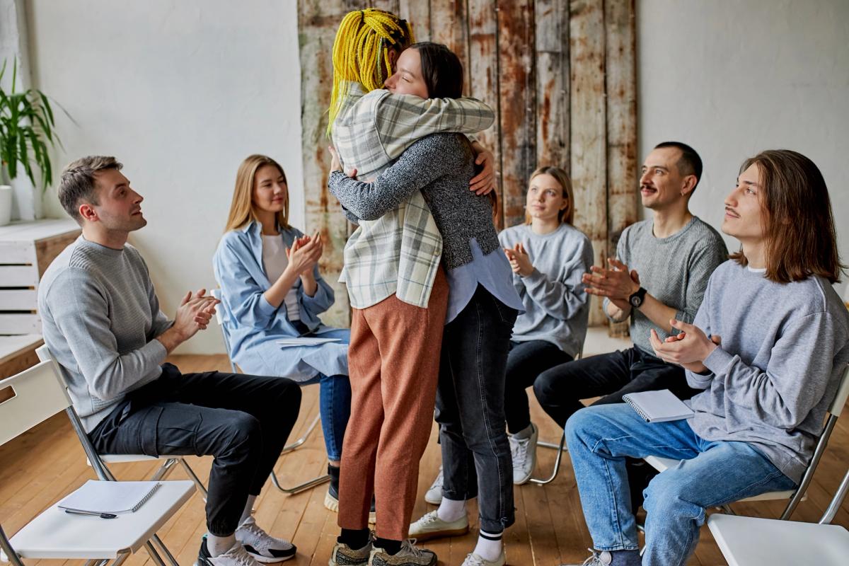Two people share a warm hug in the center of a room while a diverse group of people sitting in a circle clap and look on with supportive smiles