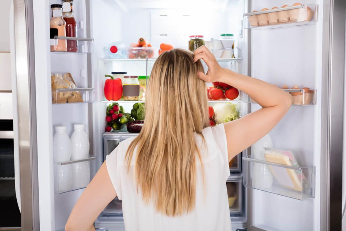 A person stands in front of an open refrigerator filled with fresh foods, scratching their head as if deciding what to choose, symbolizing moments of indecision and the challenge of avoiding self-sabotaging habits.