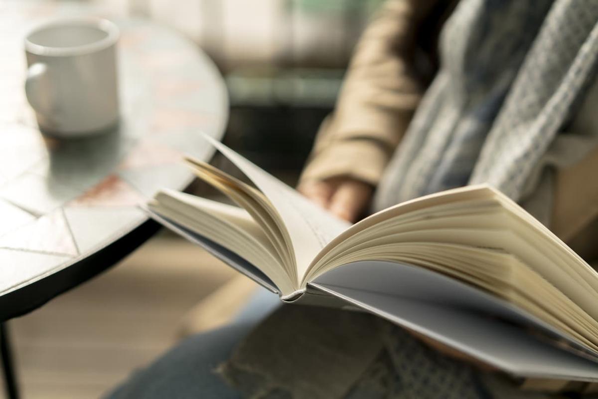 A close-up, soft-focus shot of a person's hands holding an open book, with a white coffee mug on a table in the background.