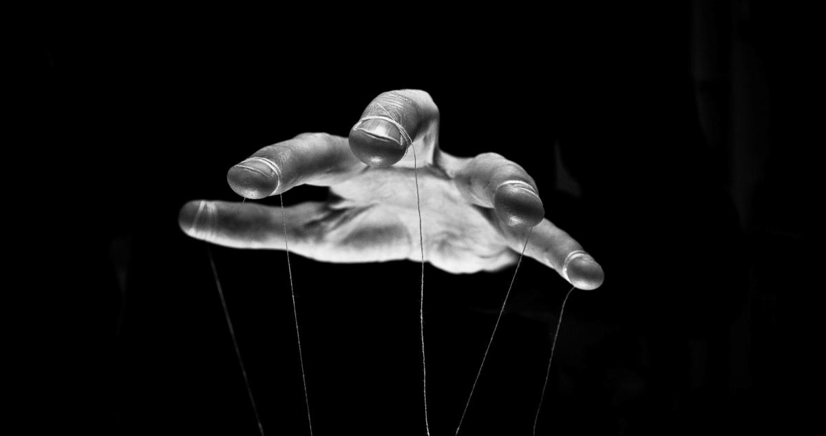 Black-and-white image of an outstretched hand holding thin strings like a puppeteer, symbolizing control, manipulation, or influence.