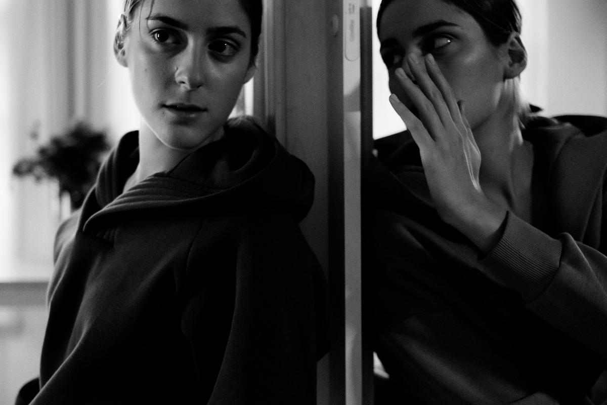 Black-and-white photo of a woman in a hoodie staring at her reflection in a mirror, with her reflected hand covering her mouth, experiencing drug-induced psychosis.
