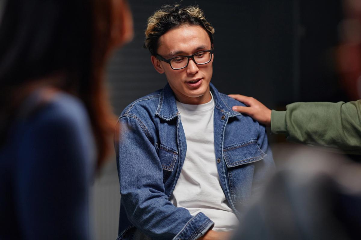 Young man speaking in a support group setting while another participant places a reassuring hand on his shoulder, representing seeking help for Valium addiction and benzodiazepine recovery.