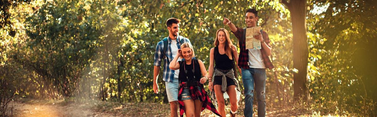 A group of four diverse young adults laughing and hiking through a sun-dappled forest. One man leads the way holding a map and pointing ahead, while the others follow closely behind on a leafy trail.