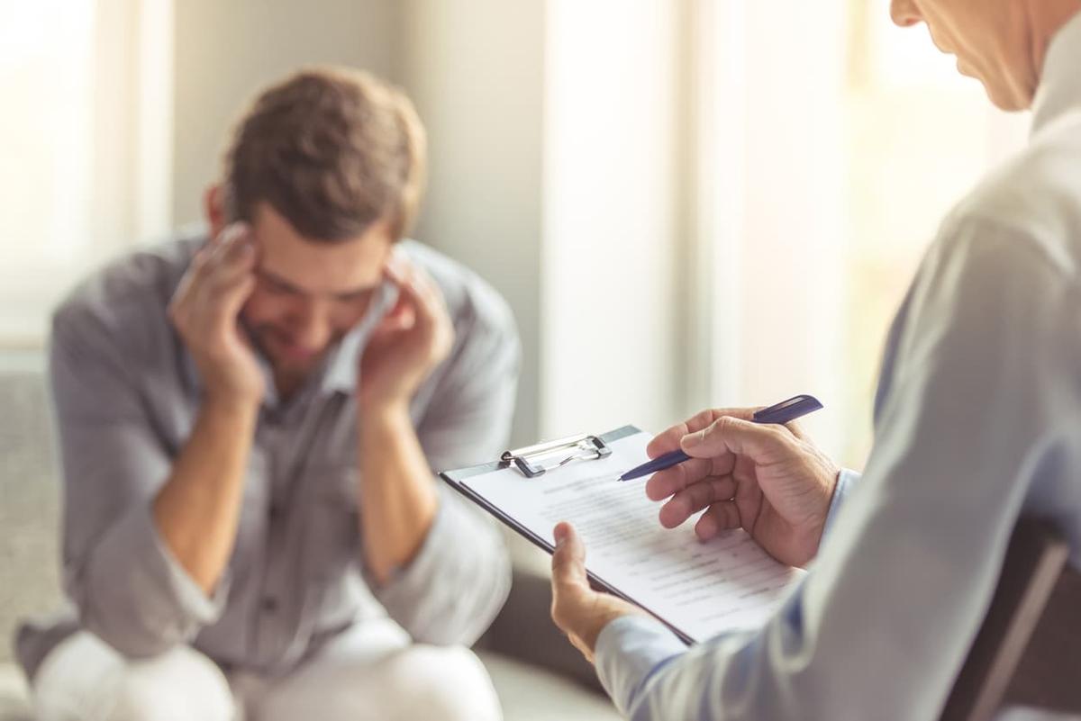 An over-the-shoulder shot of a therapist holding a clipboard and pen, looking toward a blurred man in the background who is sitting with his head in his hands, appearing distressed or thoughtful.