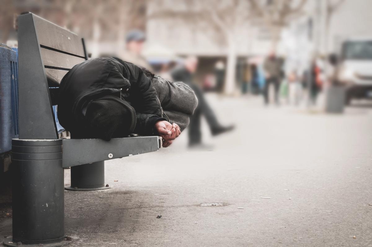 A person wearing dark, heavy clothing is curled up and sleeping on a metal and wood park bench. The background shows a blurry city walkway with figures passing by.