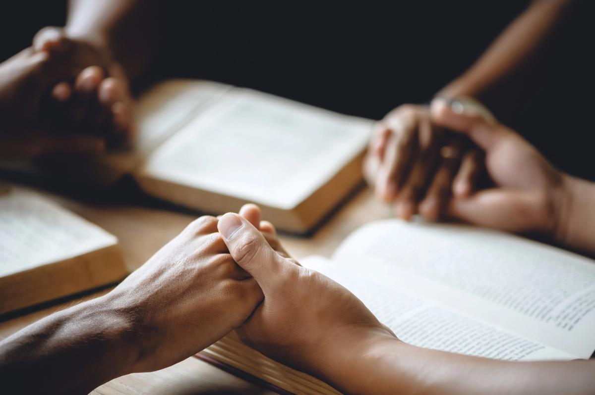 Close-up of two people holding hands across a table with open books, suggesting a group discussion or support meeting.