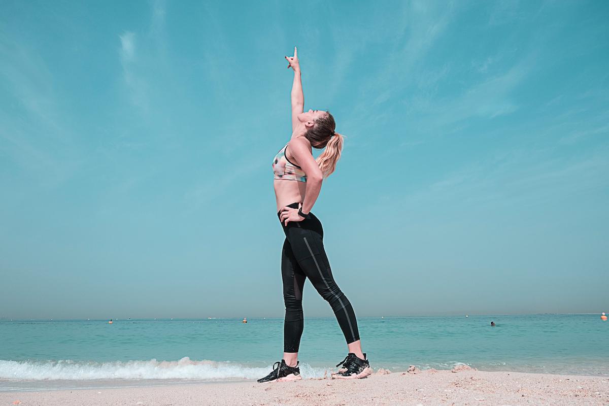 Woman in black athletic wear jumping with arms raised overhead on sandy beach with turquoise ocean and blue sky, being active in addiction recovery