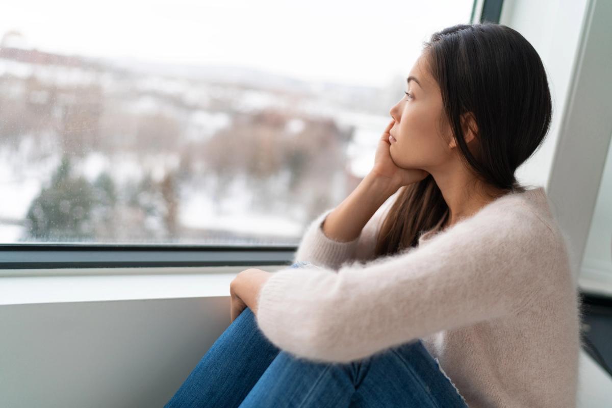 A young woman with long dark hair sits on a windowsill with her chin resting in her hand, looking thoughtfully out at a snowy, blurred landscape through a large window.