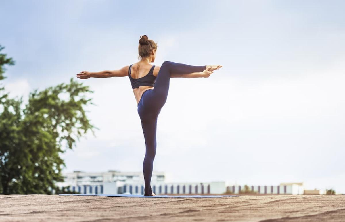 Woman practicing yoga outdoors in a standing balance pose with one leg lifted and arms extended, wearing athletic leggings and a sports bra on a rooftop or open terrace in daylight.