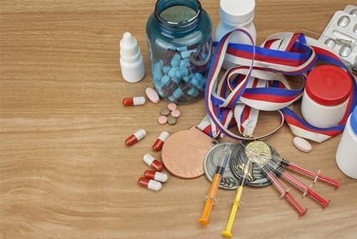Assorted pills and capsules scattered on a table beside a jar of blue tablets, prescription bottles, steroids, syringes, and several medals with red-white-blue ribbons.