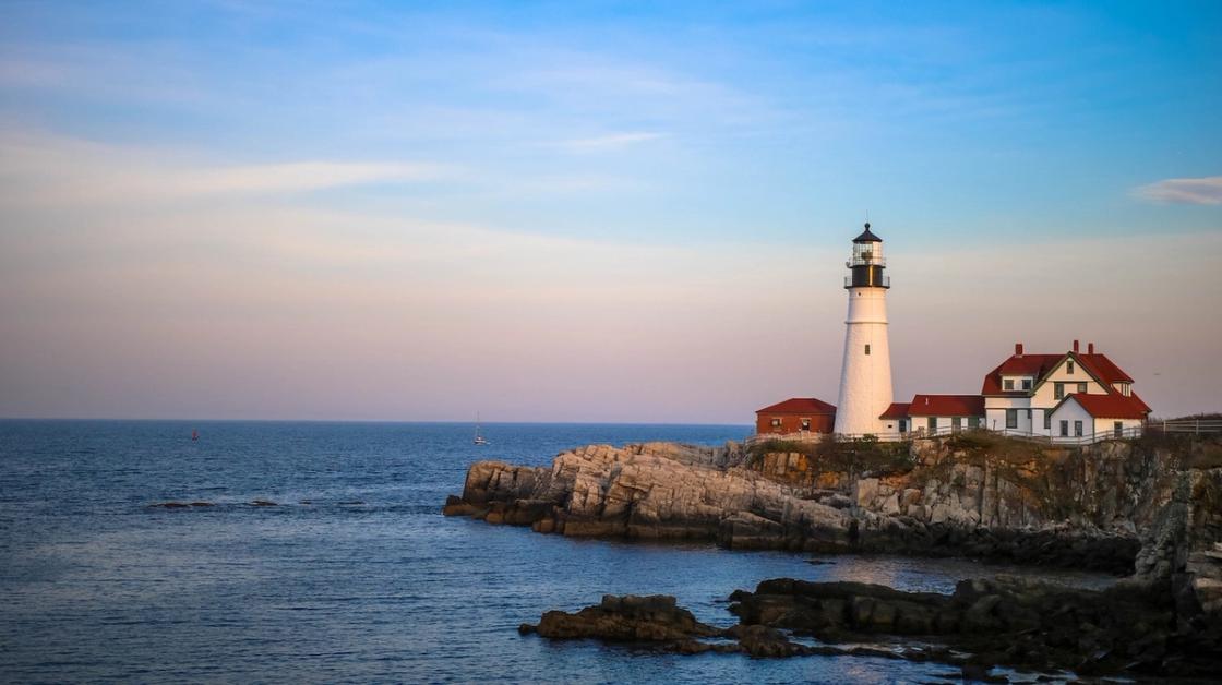 A white lighthouse and red-roofed coastal buildings sit on a rocky shoreline beside calm blue ocean waters under a pastel sky.