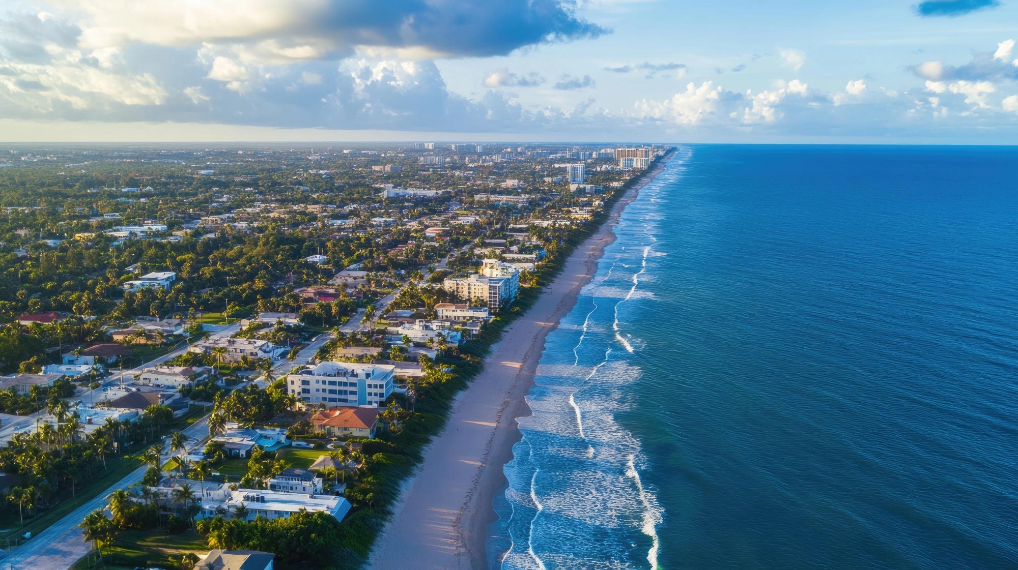 A high-altitude aerial view of a long, sandy coastline meeting bright blue ocean waves, with a lush green residential area and high-rise buildings stretching along the shore