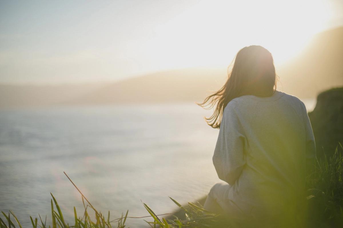 A person sits on a grassy hillside overlooking the ocean at sunrise, backlit by warm light, conveying calm reflection and resilience.