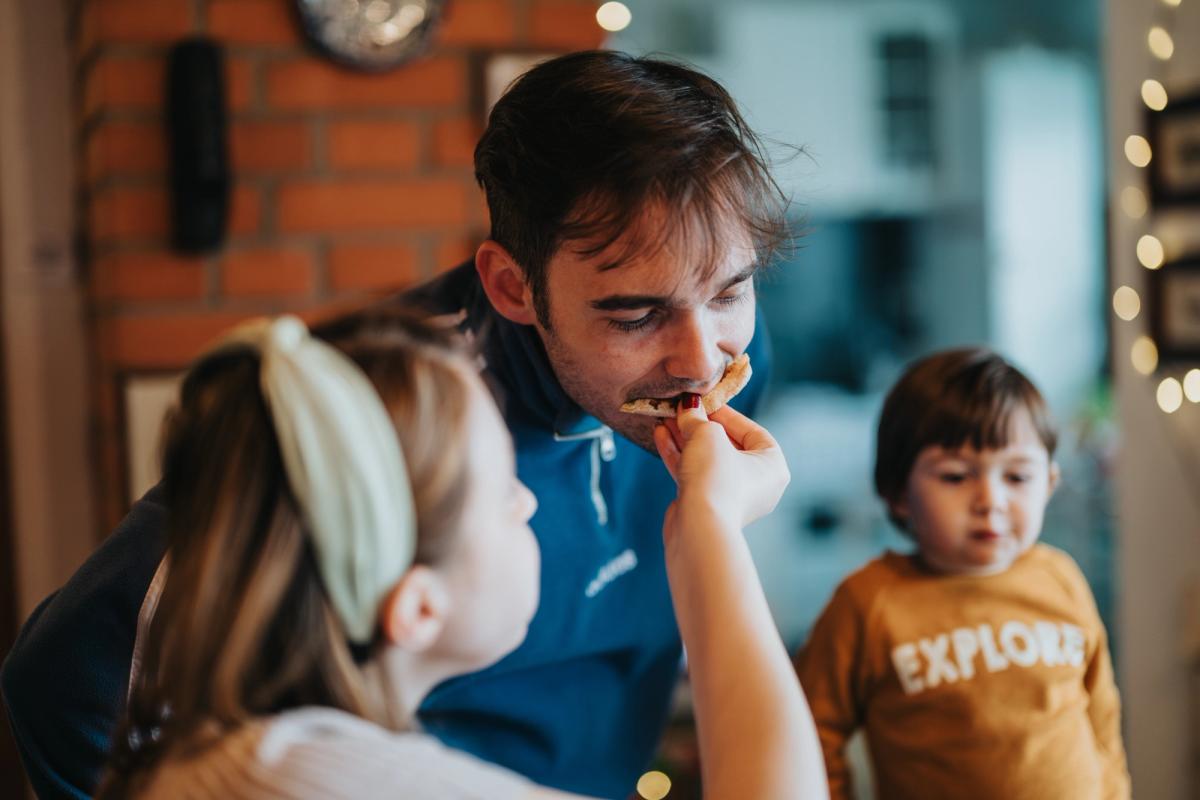 Father eating a snack while his young daughter feeds him at home, with a toddler nearby—a warm family moment in a cozy living room.