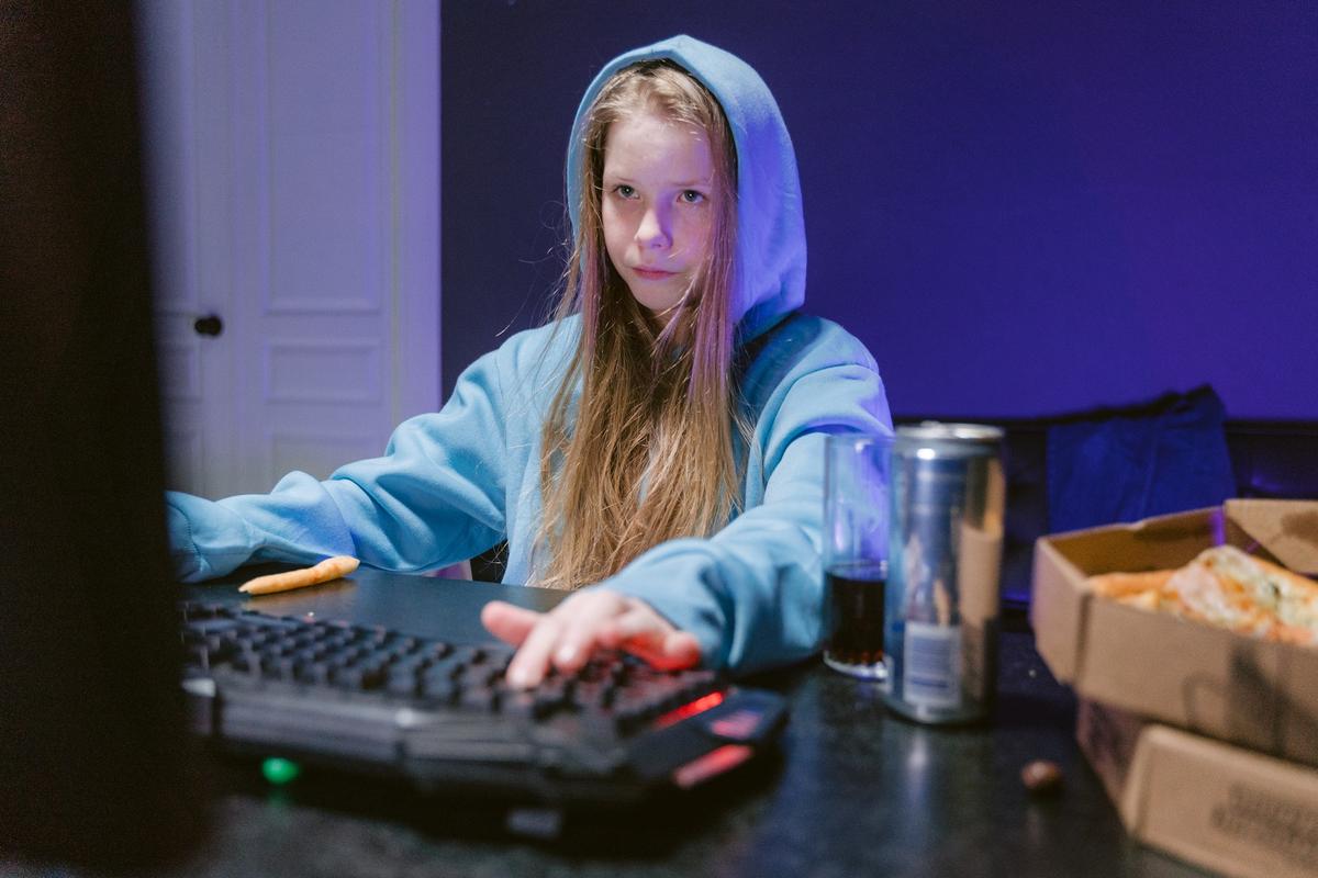 A young girl in a blue hoodie focused on a computer screen while sitting at a desk with a gaming keyboard, snacks, and a pizza box.