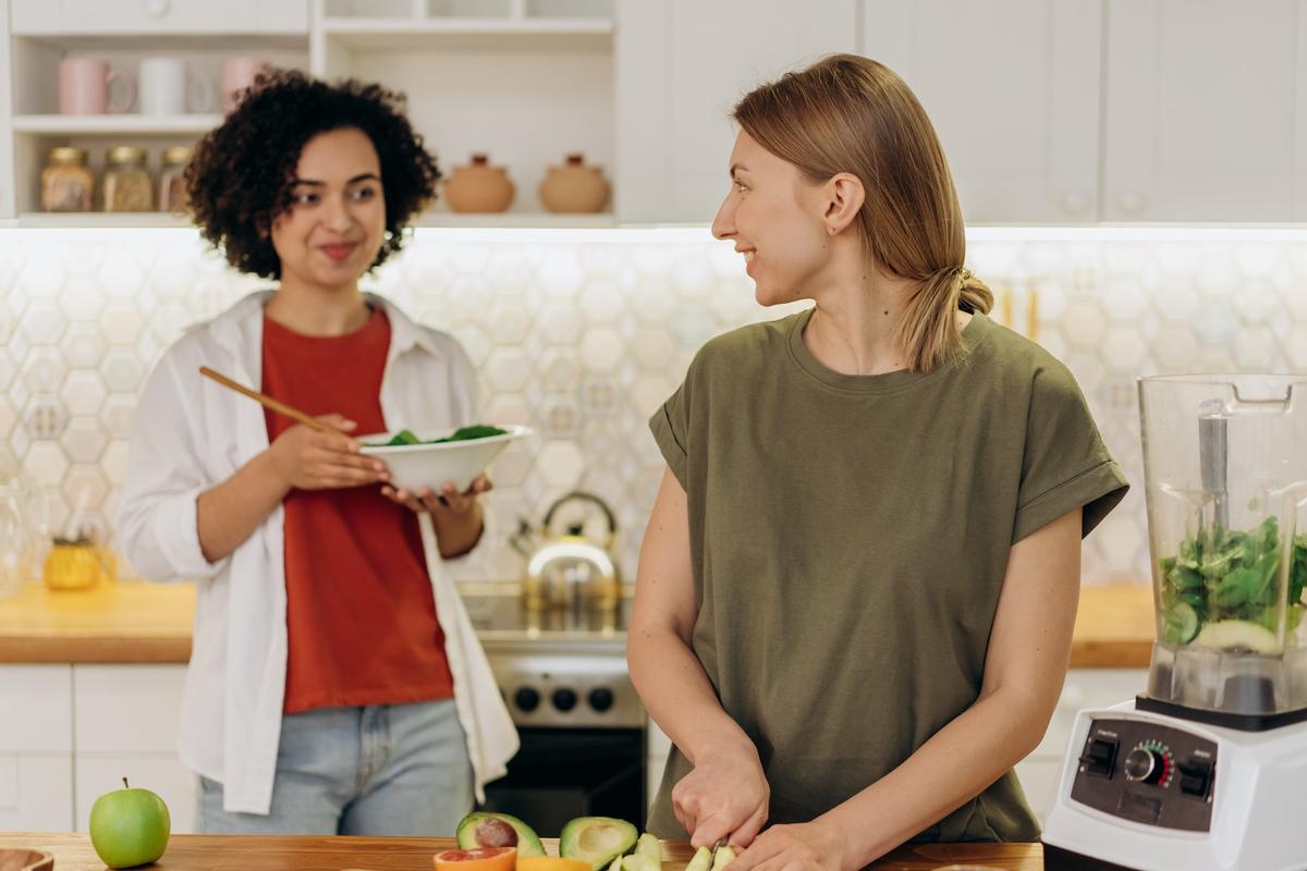 Two women smiling and talking in a bright kitchen while preparing healthy food; one chops avocados at the counter beside a blender, while the other holds a bowl of greens, suggesting positive lifestyle changes and connection during sobriety.