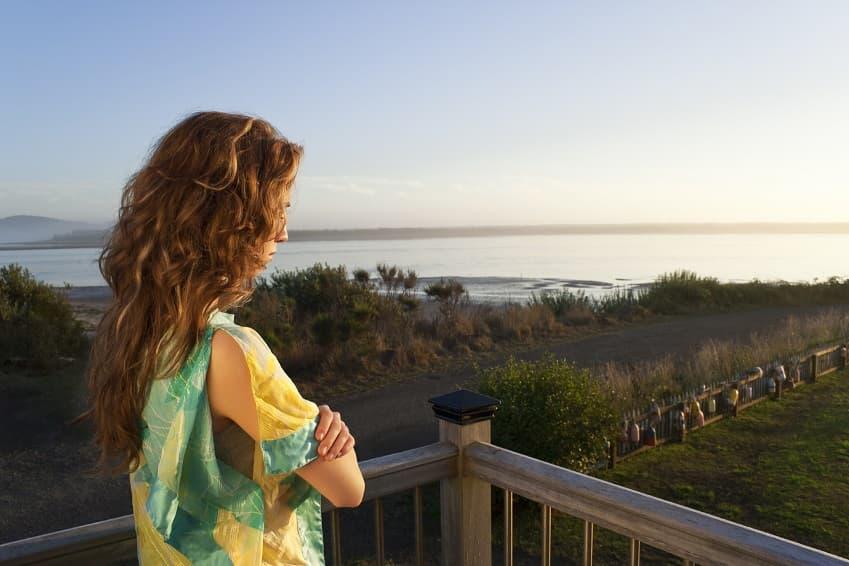 A woman with long, wavy brown hair stands on a wooden balcony with her arms crossed, looking out at a calm bay or river during sunset.
