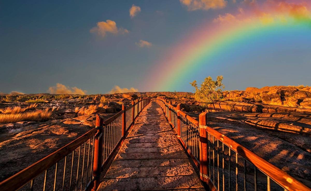A long, wooden bridge with a bright blue sky and a rainbow in the background symbolizing hope, optimism, and resilience.