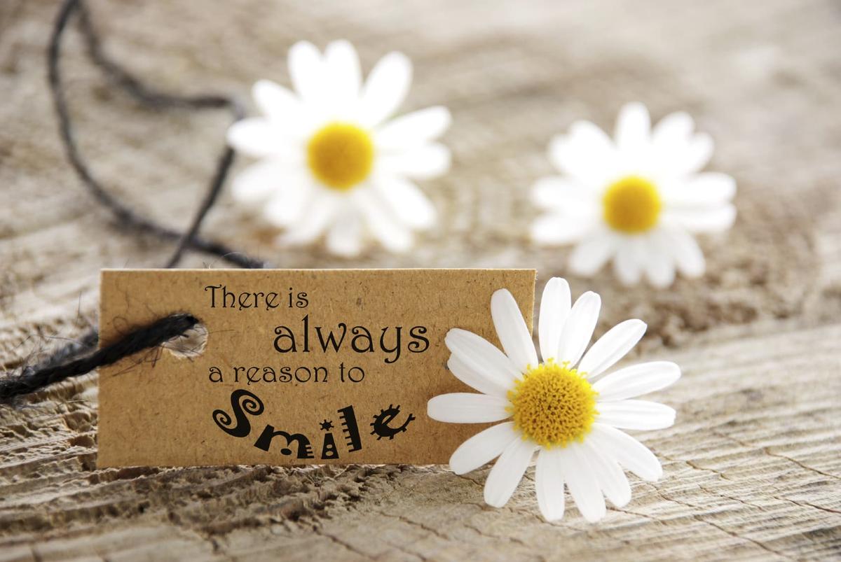 Close-up of white daisies on a rustic wooden surface beside a small brown tag tied with string that reads, “There is always a reason to smile.”