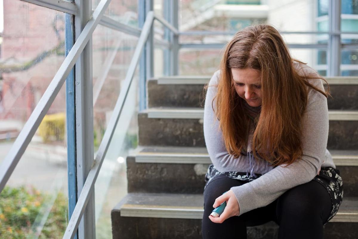 A person with chronic illness sits hunched on a stairwell landing, looking down while holding an inhaler, with glass windows and concrete steps behind them.