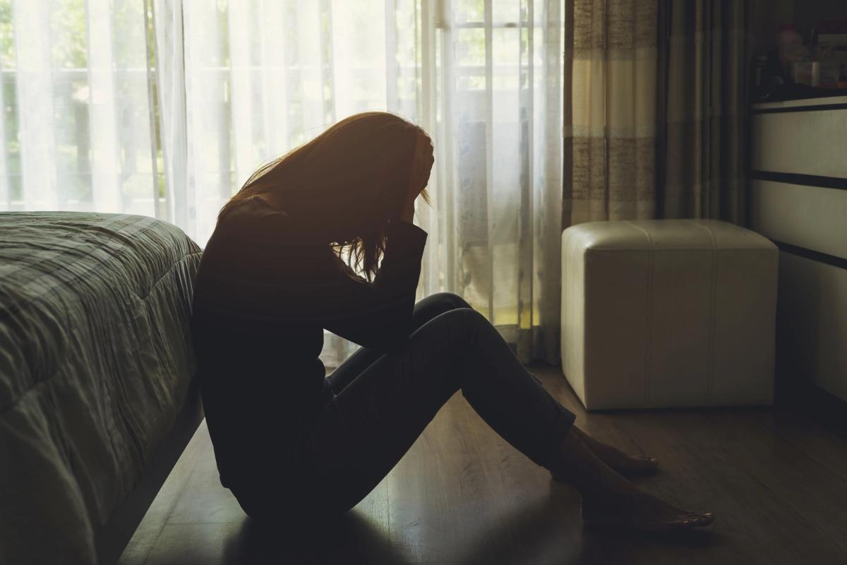 A silhouetted person sits on the floor beside a bed with their head in their hands, light filtering through sheer curtains, conveying isolation, emotional pain, and the heavy impact of untreated depression.