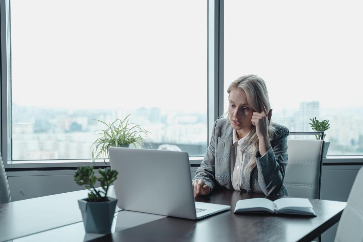 Professional woman in grey blazer sitting at office desk with laptop and open notebook, hand touching head in thoughtful or stressed gesture, large windows showing city skyline in background