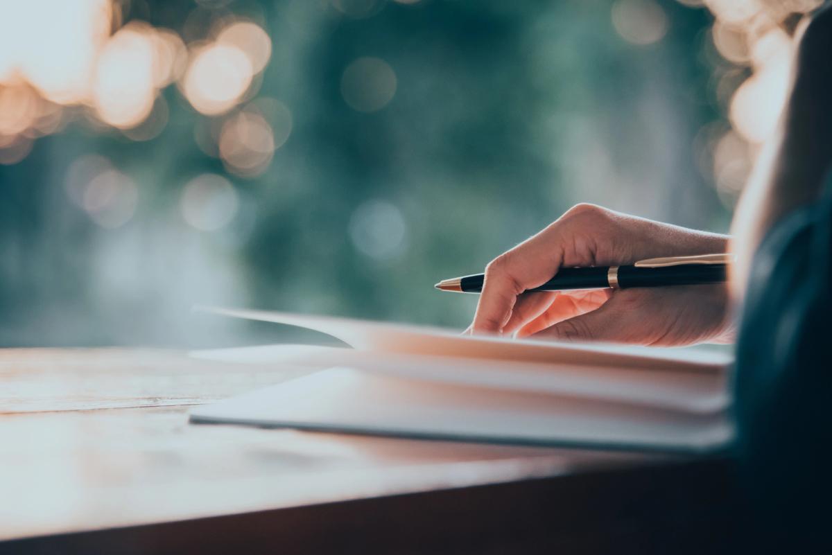 A close-up of a person's hand holding a pen over an open notebook on a wooden table.
