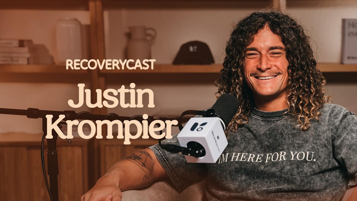 Recoverycast podcast episode featuring photographer and recovery advocate Justin Krompier smiling into a microphone while seated in a studio. Large text reads “Justin Krompier” with Recoverycast branding in the background. Justin has long curly hair, visible arm tattoos, and wears a dark gray shirt that says “I AM HERE FOR YOU.”