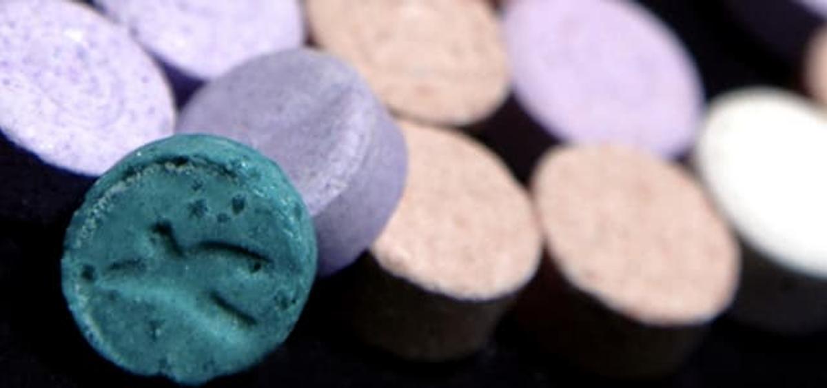 A close-up shot of several small, round, colorful pills or tablets. A teal-colored pill with an embossed bird-like logo is in sharp focus in the foreground, while other purple and beige tablets are blurred in the background.
