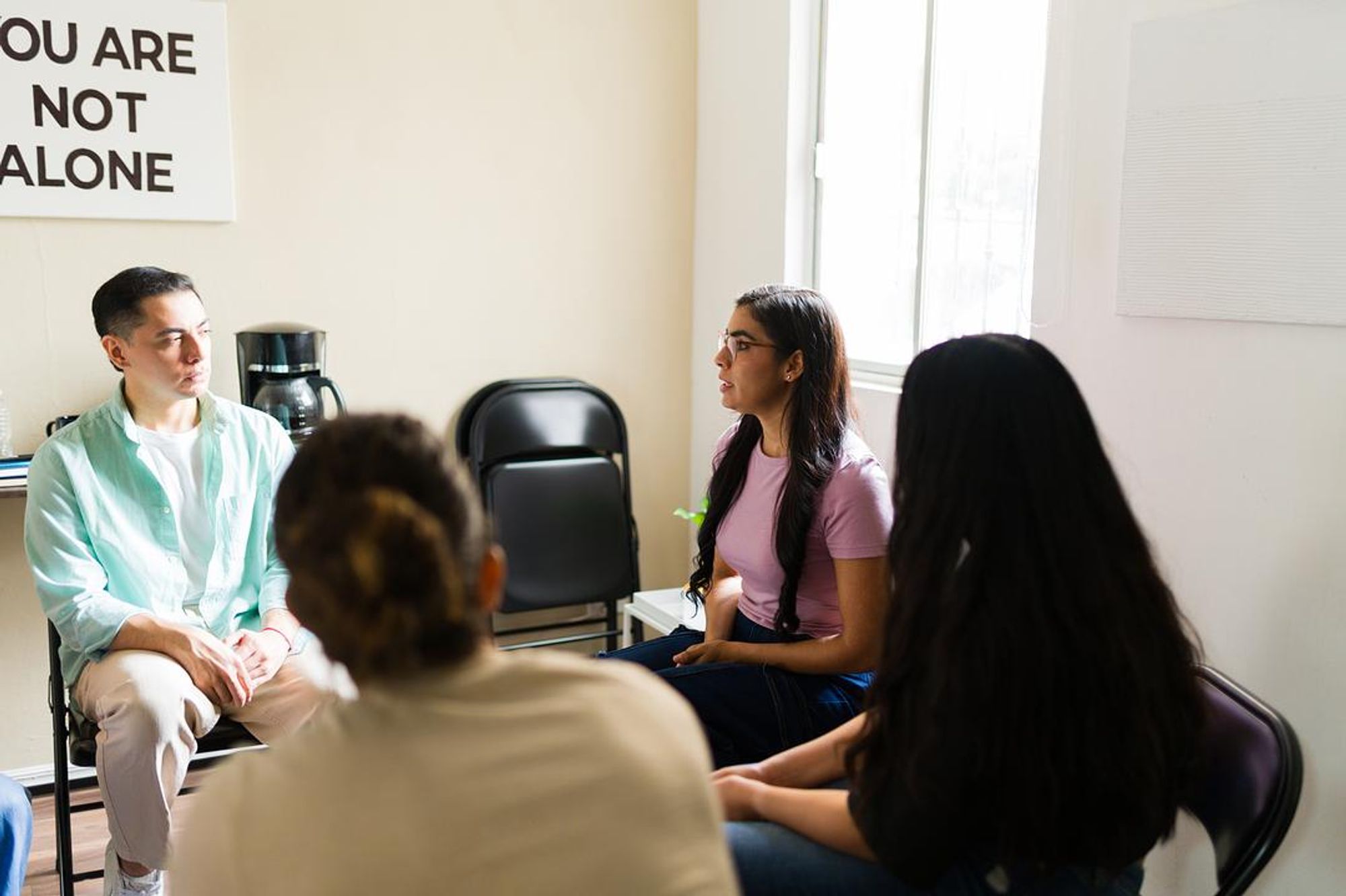 People sitting in a circle during a group therapy session at an addiction recovery support meeting with a ‘You Are Not Alone’ sign on the wall.
