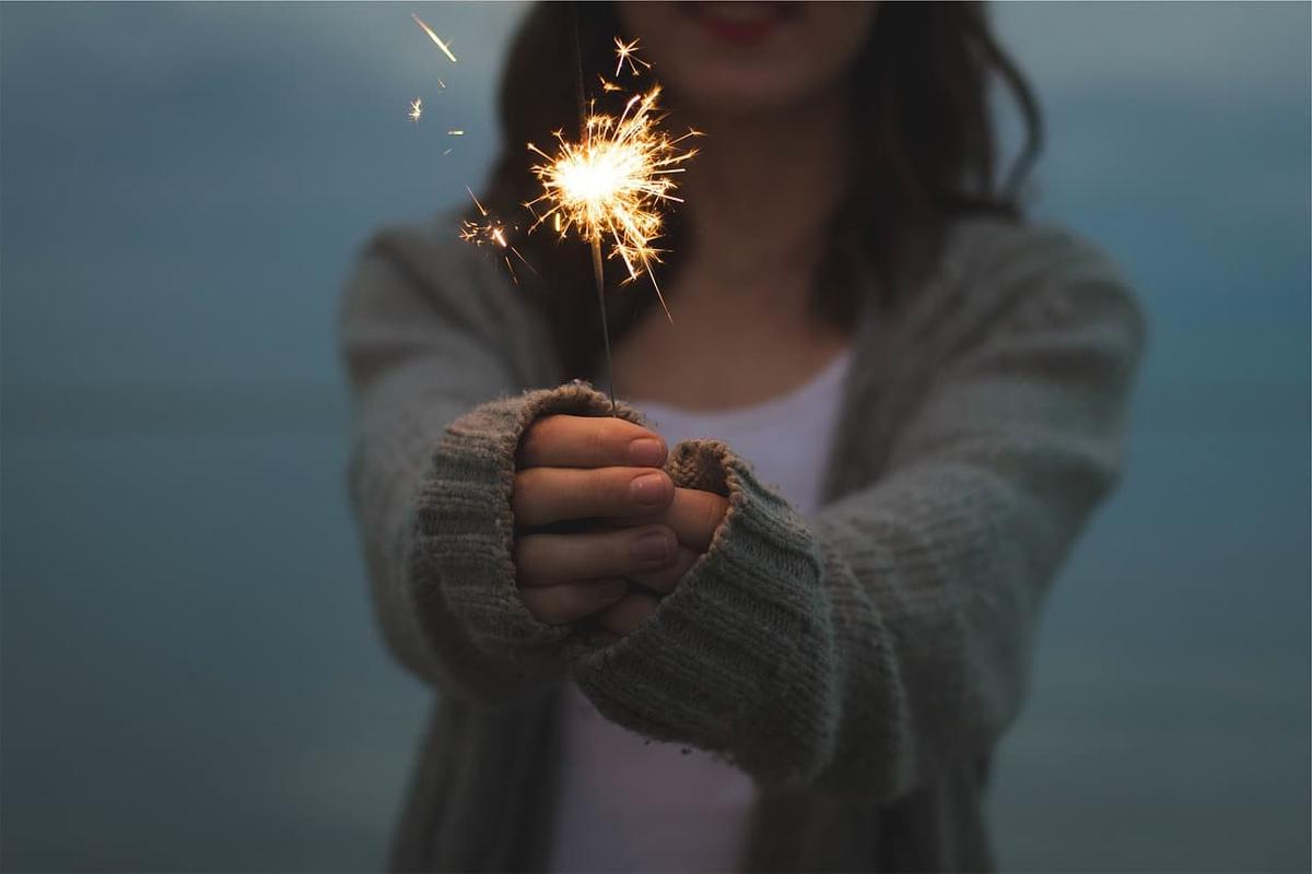 A person in a cozy sweater holds a lit sparkler with both hands, the bright sparks in focus and their face softly blurred in the background, symbolizing hope and guidance when learning how to find a recovery sponsor.