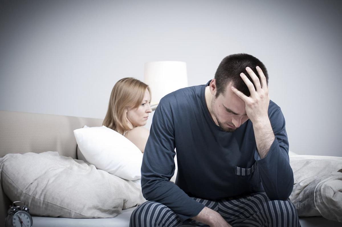 Man sitting on the edge of a bed holding his head in distress while a woman sits behind him looking concerned symbolizing relationship strain depression stress and emotional disconnect.