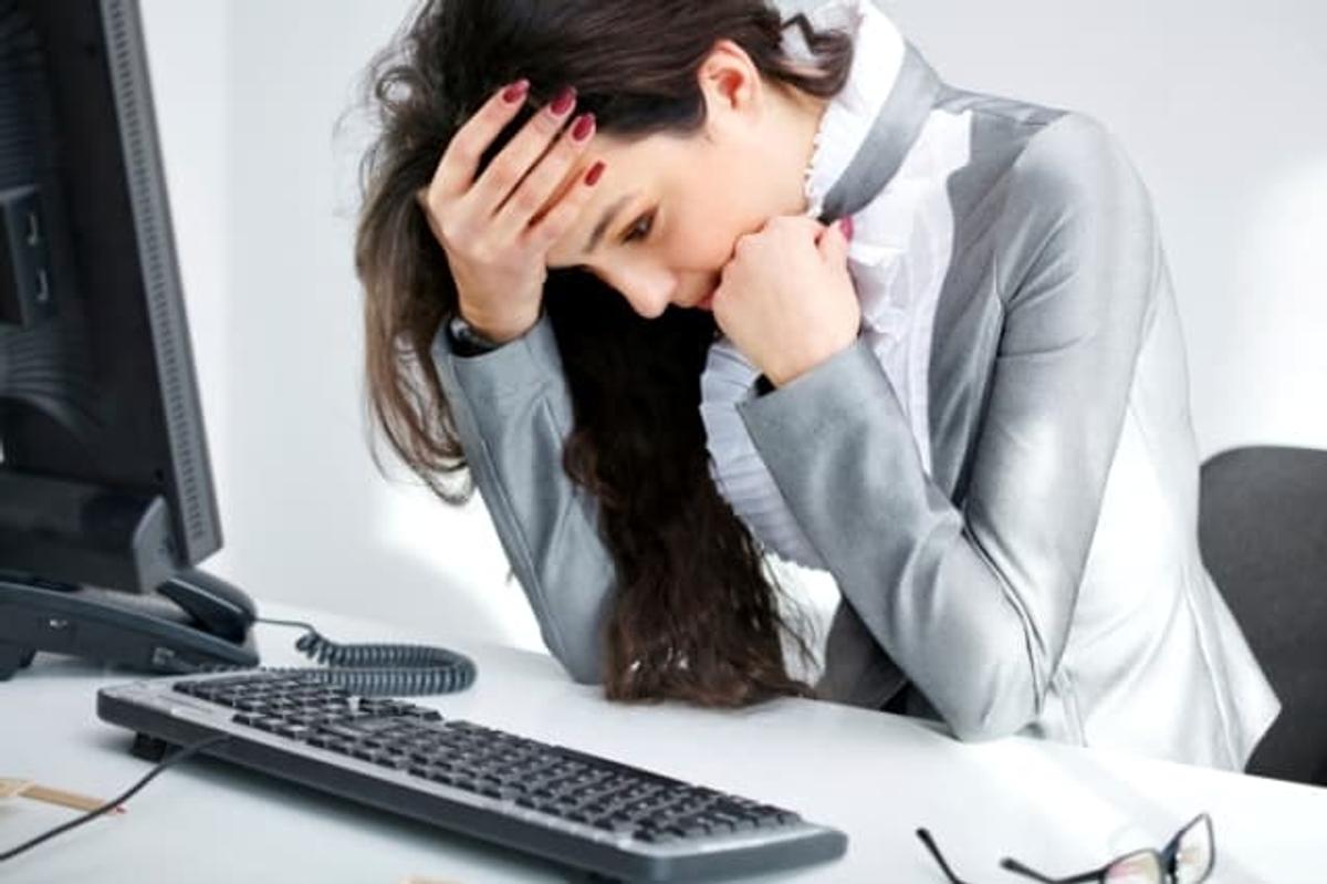 A stressed businesswoman sitting at her desk with her head in her hands, looking down at a computer keyboard