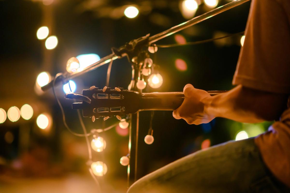 Close-up of a guitarist holding an acoustic guitar beside a microphone stand wrapped with string lights, with warm bokeh lights in the background.
