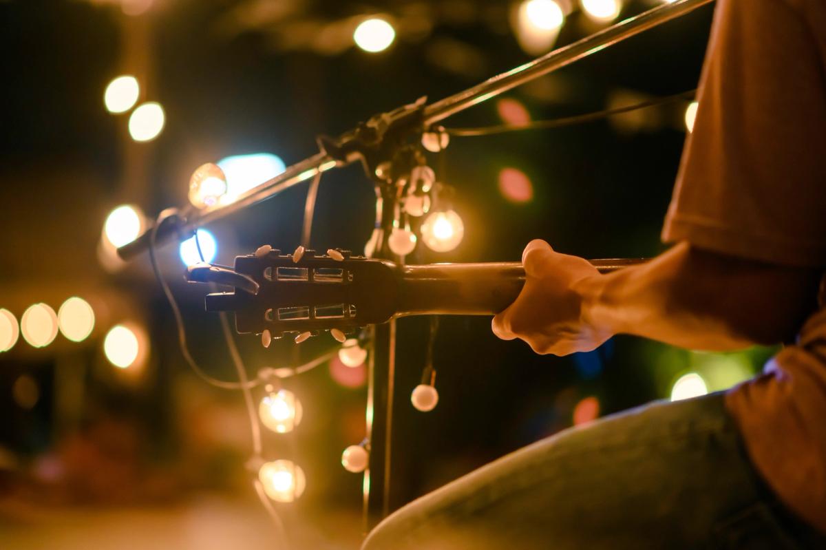 Close-up of a guitarist holding an acoustic guitar beside a microphone stand wrapped with string lights, with warm bokeh lights in the background.