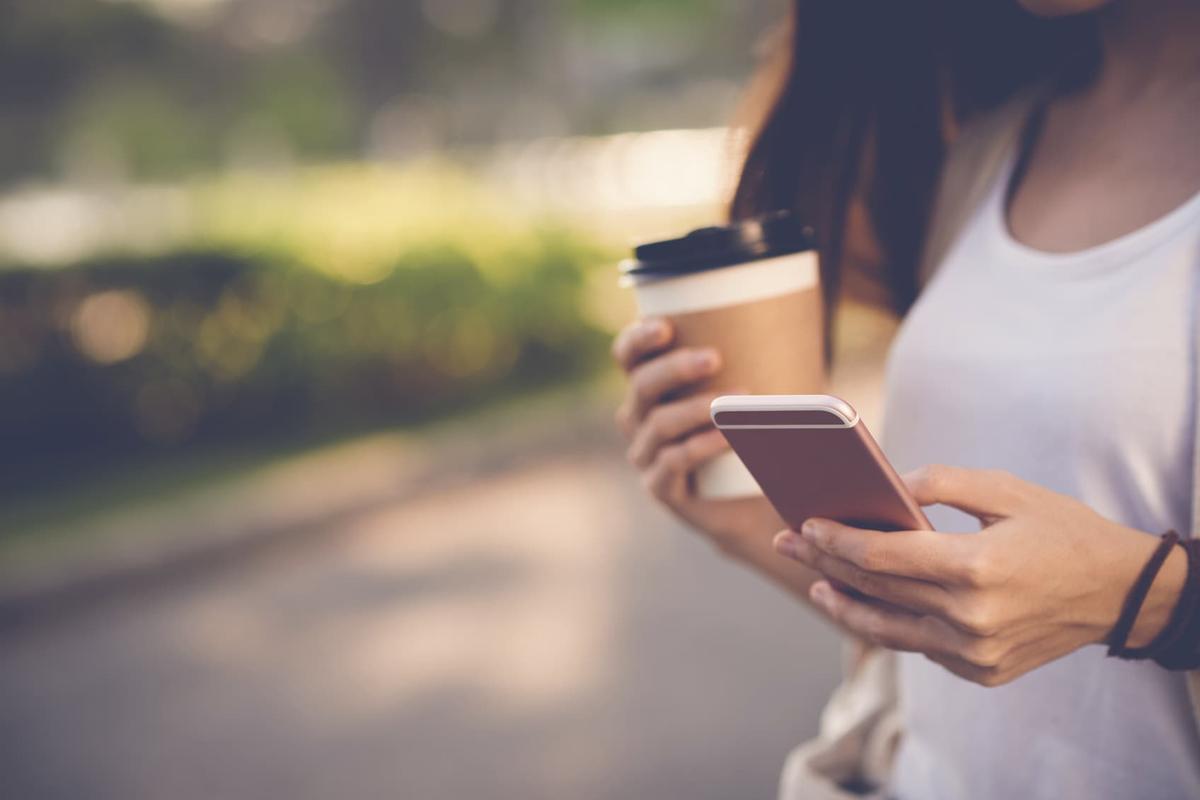 A close-up, blurred outdoor shot of a person holding a smartphone in one hand and a takeaway coffee cup in the other.