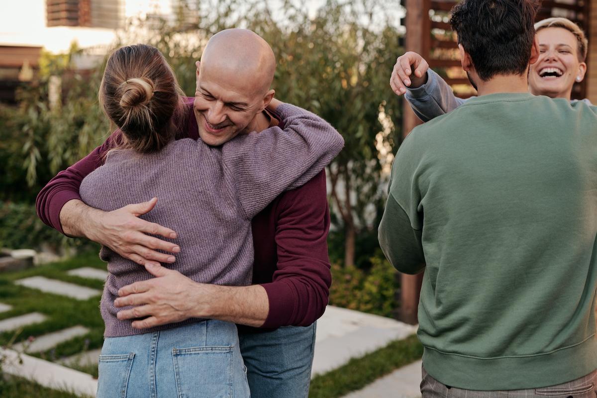 A group of four friends greeting each other with warm hugs in an outdoor garden.