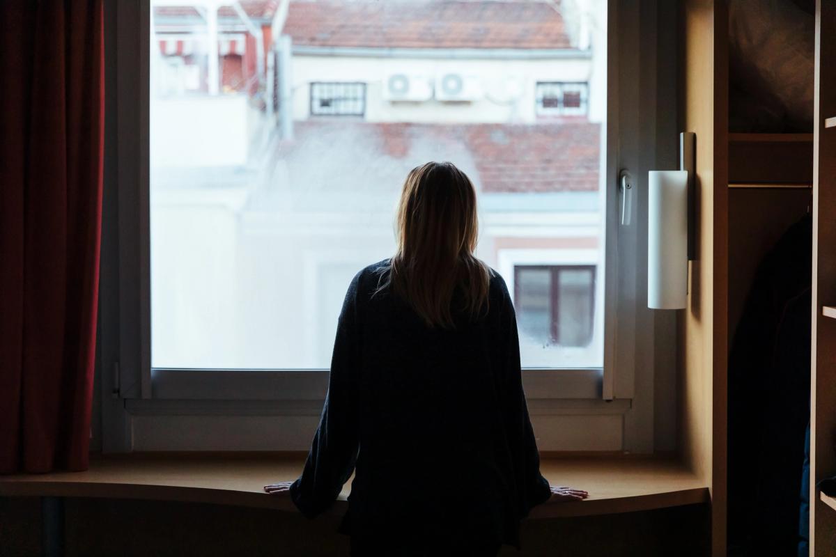 Person sitting on a windowsill indoors, looking out a large window at nearby buildings.