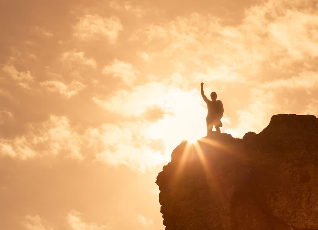 Silhouette of a person standing on a rocky cliff with one arm raised in triumph against a golden sunrise, sunbeams flaring behind them.