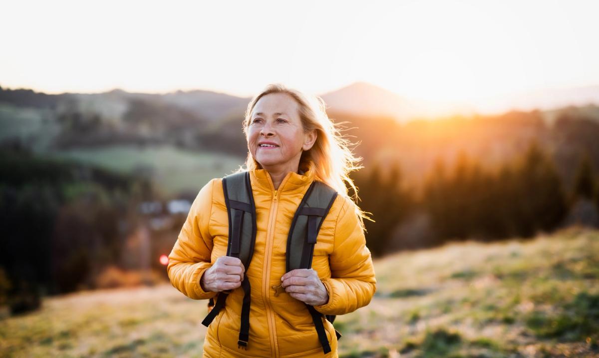 Middle-aged woman wearing a yellow jacket and backpack standing on a hillside at sunrise, looking hopeful as the morning light shines behind her—symbolizing reflection on what role your biological clock plays in recovery and new beginnings.