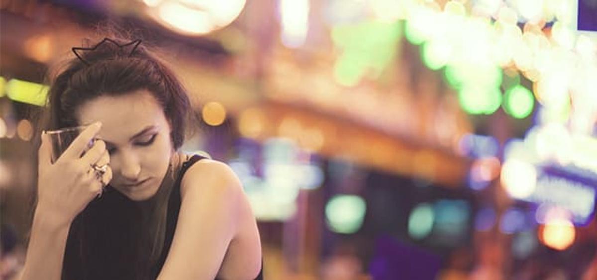 Young woman looking distressed at a bar with colorful nightlife lights in the background, illustrating alcohol misuse, emotional burnout, and the need for addiction recovery support.