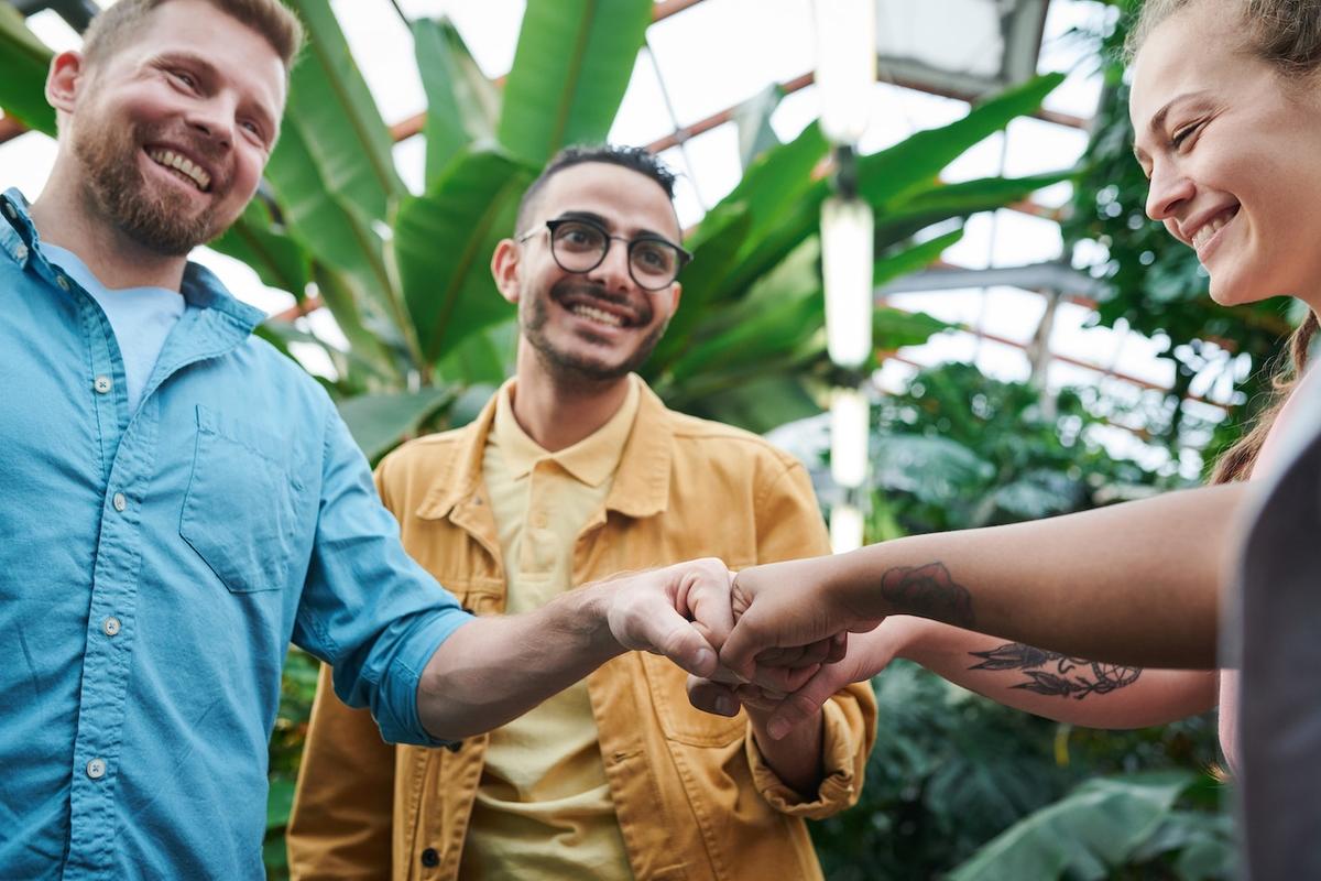 Three people in a therapeutic community doing a fist bump together while smiling, surrounded by green plants in bright indoor space, showing peer support and connection in recovery