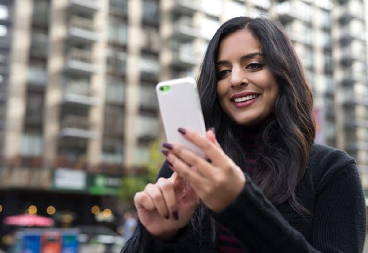 Woman smiling at her phone in an urban area, symbolizing access to online support, recovery resources, or telehealth services.