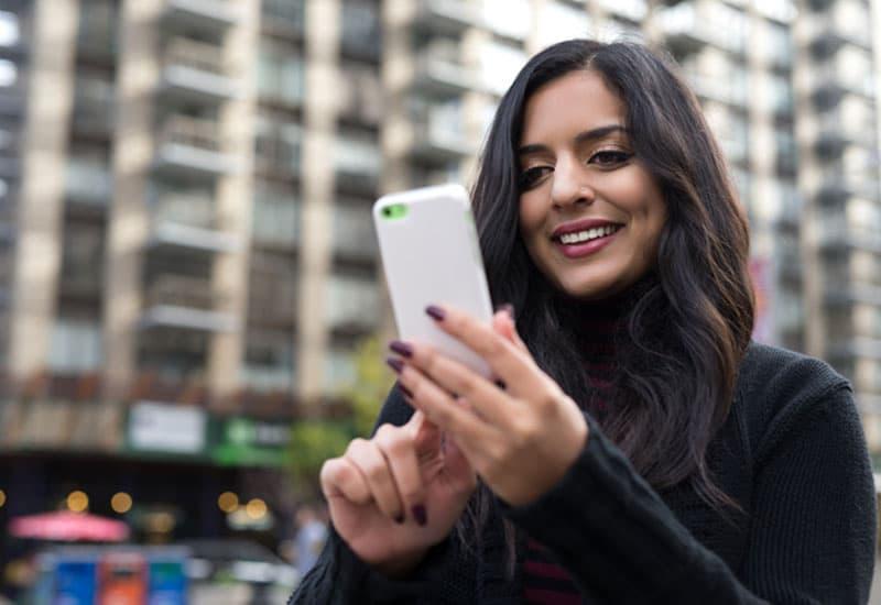 Woman smiling at her phone in an urban area, symbolizing access to online support, recovery resources, or telehealth services.