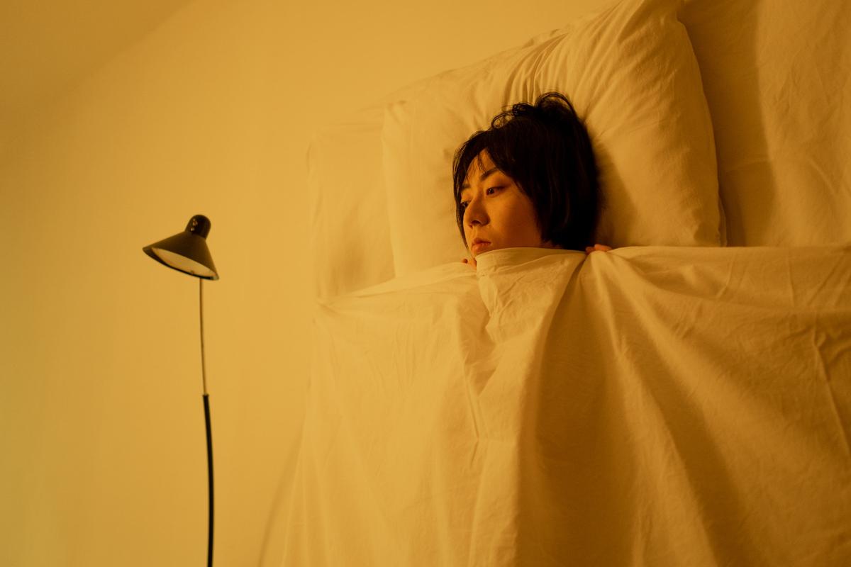 A person lying awake in bed, tucked under white sheets in a room with warm, yellow lighting.