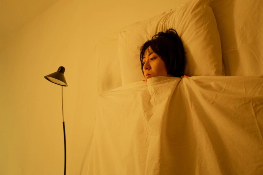 A person lying awake in bed, tucked under white sheets in a room with warm, yellow lighting.